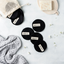 Black reusable makeup pads with visible brand label on a light gray surface with a textured fabric and small flowers.