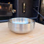 A stainless steel double wall camping bowl with the logo 'INDISPOSABLE' debossed on the front, resting on a light wood surface with a dark, blurred background.