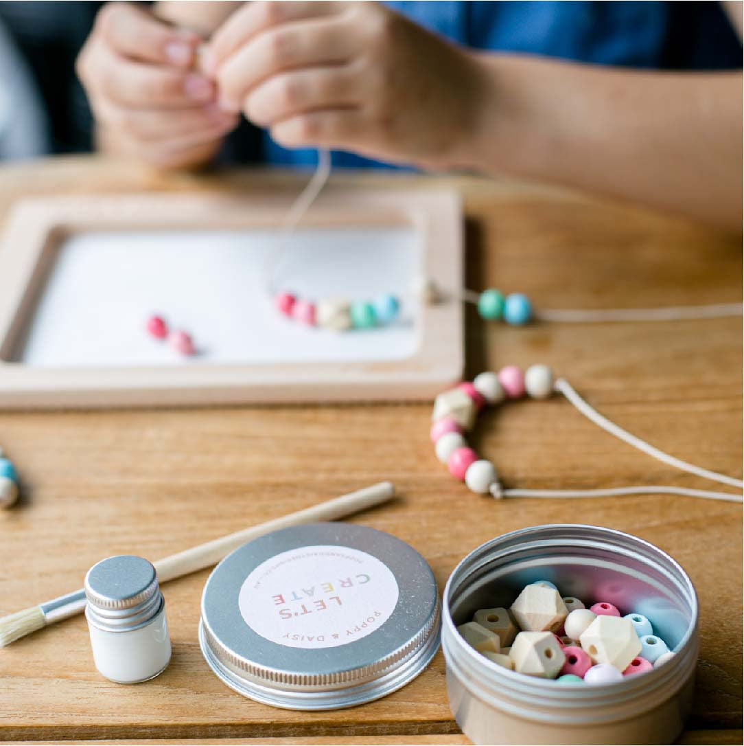 Poppy and daisy designs friendship necklace kit contents laid out showing child's hands in the background out of focus placing bead onto thread