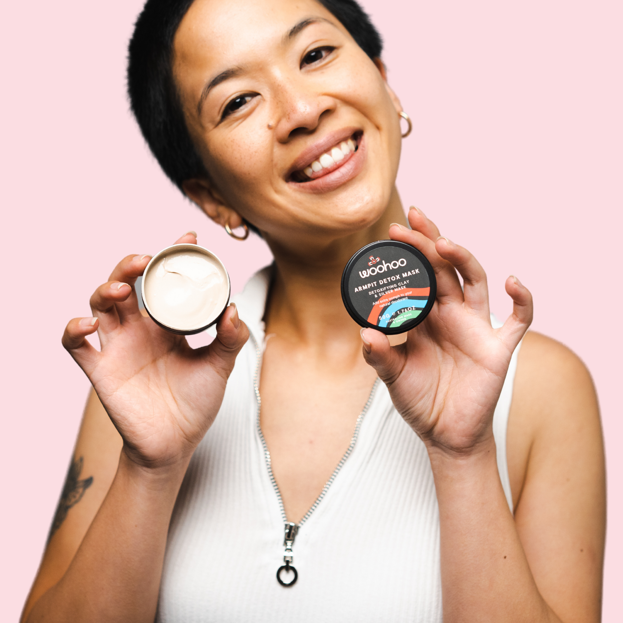 lady smiling at camera holding open tin of woohoo armpit detox mask in one hand and the tin lid in the other hand