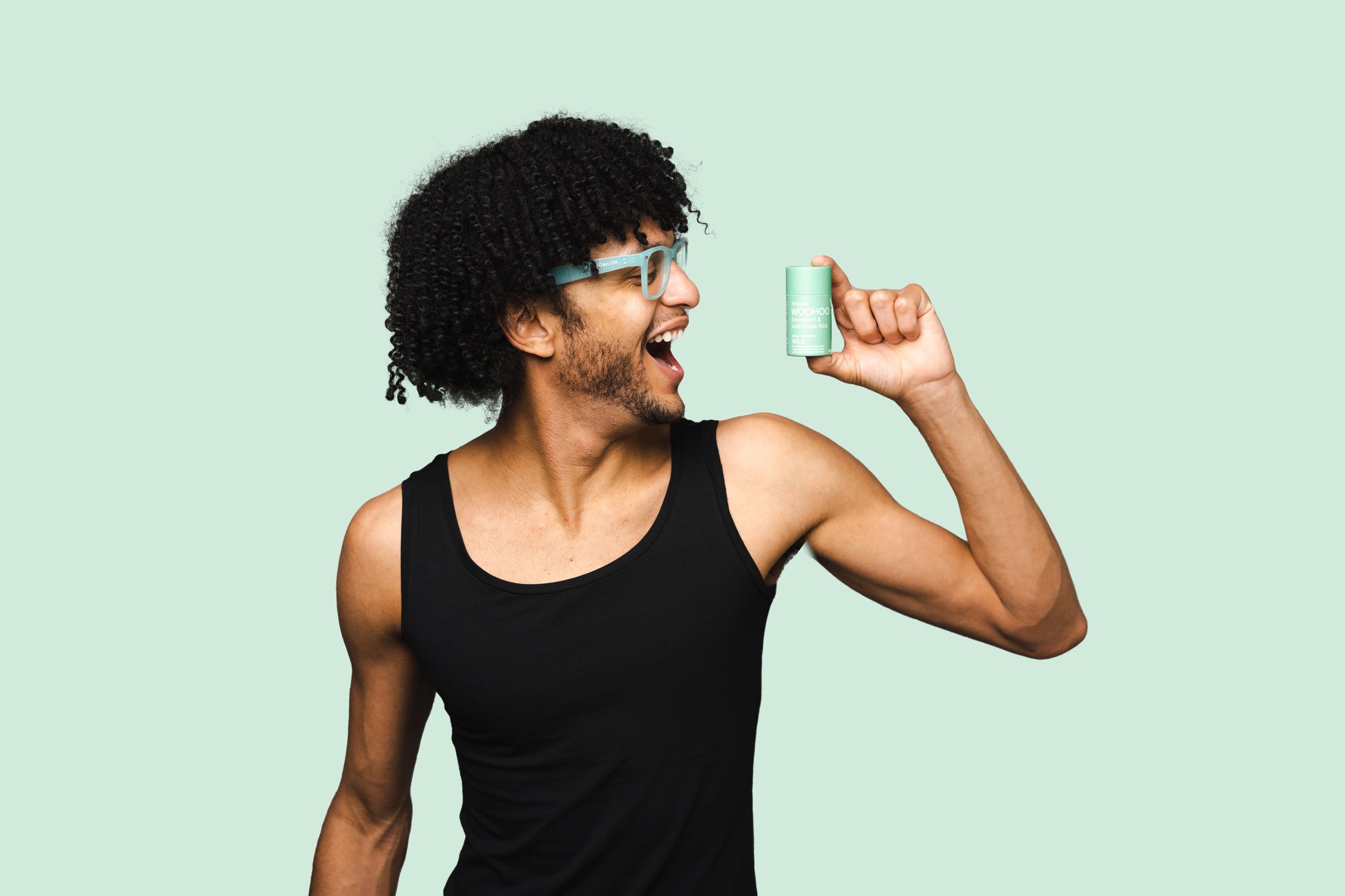man with black curly hair wearing green glasses smiling holding woohoo wild all natural deodorant and anti-chafe stick extra strength 60 grams tube